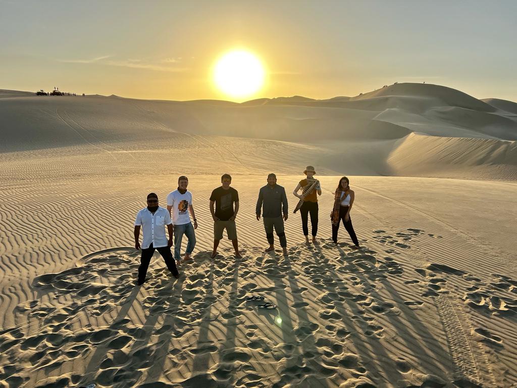 Vier Männer und zwei Frauen stehen in der Wüste. HInter ihnen sieht man eine Dünenlandschaft und die untergehende Sonne.