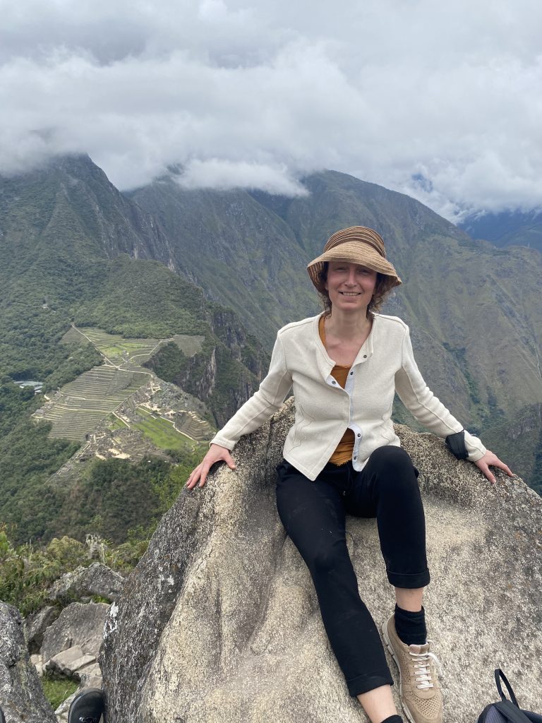 Ich, eine Frau sitze auf einem Stein. Hinter mir ist Macchu Picchu in Peru. Ich trage einen Sonnenhut und lache.